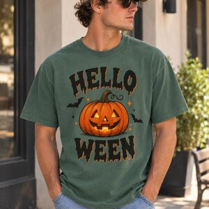 Könnte beinhalten: Ein salbeigrünes Kurzarm-T-Shirt mit einer Grafik eines leuchtenden orangefarbenen Kürbis-Gesichts. Der Text "HELLO" steht über dem Kürbis und "WEEN" darunter in einer schwarzen, tropfenden Schriftart, ergänzt durch kleine schwarze Fledermäuse.