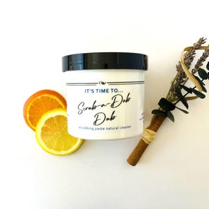 May include: A white cylindrical container with a black lid, labeled with the text IT'S TIME TO... Scrub-a-Dub Dub scrubbing paste natural cleaner. Beside the container are two citrus slices and a bundle of dried lavender and eucalyptus.