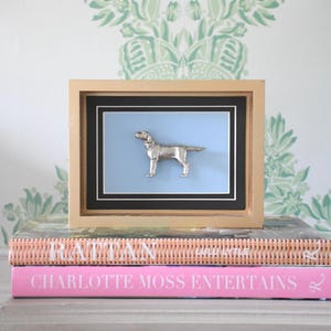 May include: A framed wall art piece featuring a three-dimensional silver-toned dog figurine mounted on a light blue background. The artwork is enclosed in a rectangular gold-colored frame with a black and white matting border surrounding the dog.