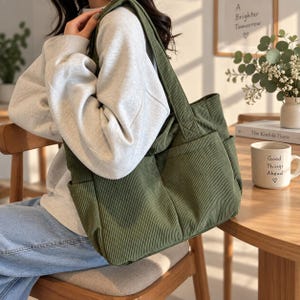 Op de afbeelding: Een olijfgroene corduroy tote bag met een geribbelde textuur, twee brede schouderbanden en twee grote buitenzakken aan de voorzijde. De tas heeft een rechthoekige vorm met een ruim hoofdvak en zichtbare verticale stiksels.
