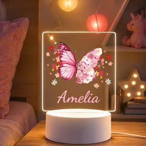 Könnte beinhalten: Ein rechteckiges LED-Nachtlicht mit weißem, rundem Sockel. Die klare Acrylplatte zeigt einen aufgedruckten Schmetterling mit einem rosa Flügel und einem Flügel aus rosa Blüten, umgeben von kleinen Blumenmotiven und dem Namen Amelia in rosa Schrift.