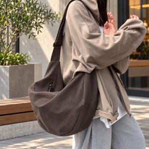 May include: A dark brown hobo-style shoulder bag made of a soft, matte fabric. The bag features a wide, adjustable shoulder strap and a visible vertical zipper pocket on the front exterior. The design is slouchy with a curved, crescent-like shape.