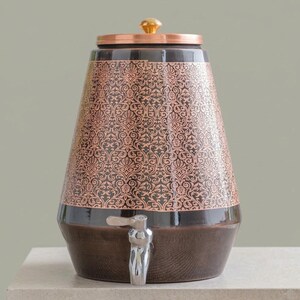 Puede incluir: Un dispensador de bebidas cónico con una base marrón oscuro y una sección superior de color cobre con un intrincado patrón floral repetitivo. El recipiente incluye una tapa metálica con un pequeño pomo y un grifo plateado en la parte frontal.
