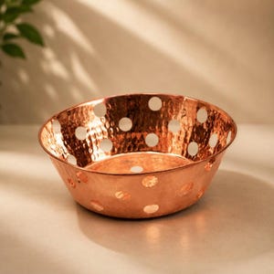 Puede incluir: Un cuenco redondo de cobre martillado que presenta una serie de recortes circulares espaciados uniformemente por todo su cuerpo. La superficie metálica tiene un tono cálido marrón rojizo con un acabado reflectante y una textura marcada.