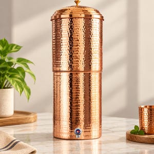 May include: A tall, cylindrical copper water dispenser featuring a hammered texture throughout its exterior. The vessel includes a removable lid with a small knob handle and a silver-toned push-button spigot located at the base for dispensing liquid.