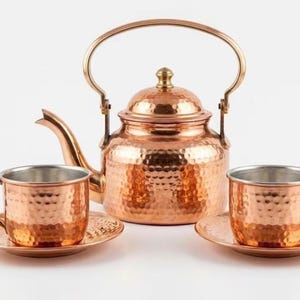 May include: A hammered copper tea set consisting of a teapot with a curved spout and arched handle, accompanied by two matching cups and saucers. Each piece features a textured, dimpled surface and a polished metallic finish in warm copper tones.