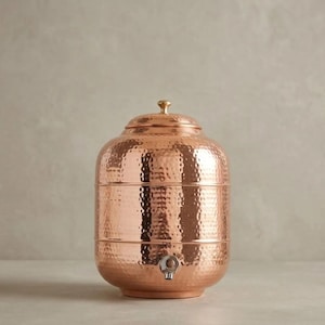 May include: A cylindrical copper water dispenser featuring a hammered texture across its surface. The vessel includes a matching lid with a small knob handle at the top and a silver-toned metal spigot positioned near the base for dispensing liquid.