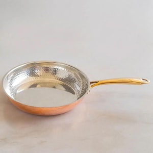 May include: A round frying pan with a hammered copper exterior and a polished silver-toned interior. The pan features a long, smooth, gold-toned metal handle attached to the side, extending outward with a small circular hole at the very end.