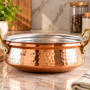 May include: A round, hammered copper serving bowl with a metallic silver-toned interior and two polished gold-toned metal handles attached to the sides. The exterior surface features a textured, dimpled pattern across the warm copper body.