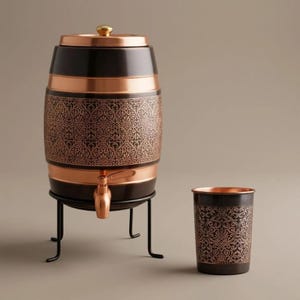 May include: A barrel-shaped beverage dispenser with a copper-coloured lid, base, and spigot, featuring a dark brown body with an intricate copper-toned damask pattern. It rests on a black metal stand, accompanied by a matching dark brown and copper cup.