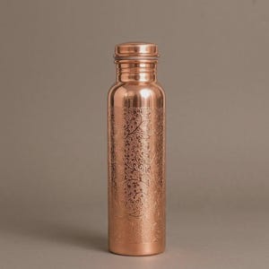 Könnte beinhalten: Eine zylindrische Wasserflasche aus Kupfer mit polierter Metalloberfläche und einem detaillierten, filigranen Blumenmuster, das in den unteren Teil eingraviert ist. Die Flasche hat einen glatten Hals und einen passenden Schraubverschluss.