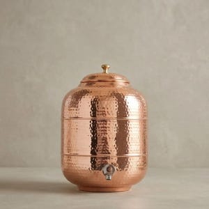 May include: A cylindrical copper water dispenser featuring a hammered texture across its surface. The vessel includes a matching lid with a small knob handle at the top and a silver-toned metal spigot positioned near the base for dispensing liquid.