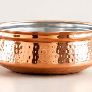 May include: A round, wide-mouthed serving bowl featuring a polished copper exterior with a distinct hammered texture. The interior of the bowl is lined with a smooth, reflective silver-coloured metal, and the rim is slightly flared and rounded.