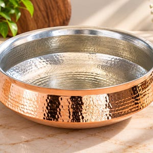 May include: A round serving bowl featuring a hammered copper exterior and a smooth, polished silver-toned interior. The bowl has a wide, flared rim and a rounded base, showcasing a distinct contrast between the warm metallic copper and the cool silver finish.