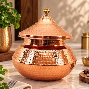 May include: A round, hammered copper pot with a wide base and a matching lid. The lid features a polished gold-tone metal knob at the center. The exterior surface of both the pot and the lid has a textured, dimpled finish that reflects light.
