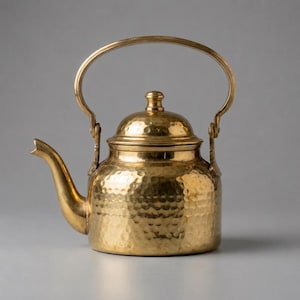 May include: A polished brass teapot featuring a distinct hammered texture across its rounded body. The vessel includes a curved spout, a matching lid with a small knob handle, and a tall, arched swing handle attached to the sides with metal brackets.