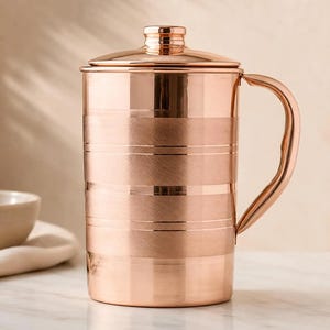 May include: A cylindrical copper pitcher with a polished finish, featuring a lid with a small knob handle and a curved side handle. The body of the pitcher displays alternating bands of brushed and smooth textures, creating a striped appearance.