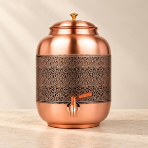 Puede incluir: Un dispensador de agua cilíndrico de cobre que presenta una banda central ancha con un intrincado patrón de filigrana negra. El recipiente tiene un acabado metálico liso, una tapa extraíble con un pequeño pomo dorado y un grifo de color cobre.