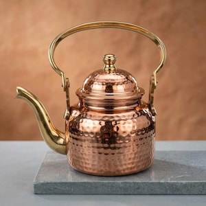 May include: A hammered copper tea kettle featuring a polished brass handle, a matching brass spout, and a domed lid with a small brass knob. The body of the kettle has a distinct dimpled texture, reflecting light across its metallic surface.