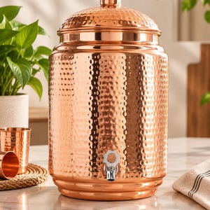 Puede incluir: Un dispensador de agua cilíndrico de cobre con una textura martillada en todo el cuerpo y una tapa abovedada a juego. El recipiente incluye un grifo de metal plateado colocado cerca de la base para dispensar líquido, con un acabado pulido.