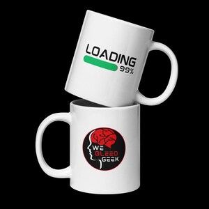 May include: Two white ceramic mugs stacked vertically. The top mug features the text LOADING with a green progress bar and 99%. The bottom mug displays a circular logo with a red brain silhouette and the text WE BLEED GEEK in black and red.