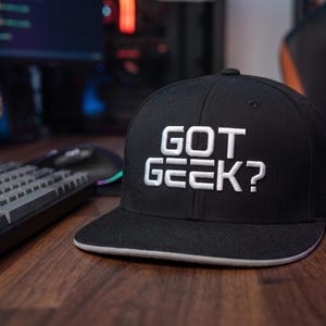 以下が含まれることがあります： 前面にGOT GEEK?と白い刺繍が施された、フラットなつばを持つ黒いベースボールキャップ。キャップは構造化されたクラウンと、つばの端に沿った白いトリムが特徴で、大胆なレタリングが施されたクラシックなデザインです。