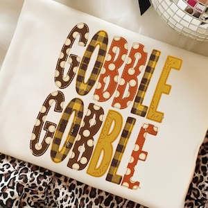 May include: A cream-colored sweatshirt featuring the text GOBBLE GOBBLE printed in two rows. The letters are decorated with various patterns including brown polka dots, yellow and black plaid, and solid orange, creating a festive autumn design.