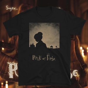 Può includere: Una t-shirt nera a maniche corte con una grafica quadrata centrale. La grafica raffigura la sagoma scura di una persona con un'acconciatura a chignon su uno sfondo strutturato e tenue, con il testo Rule of Rose stampato sotto in un carattere stilizzato.