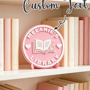 Puede incluir: Un letrero circular rosa con borde blanco que presenta el texto blanco en relieve "MELANIE'S LIBRARY" y un gráfico central de un libro abierto con un bolígrafo. Dos pequeños corazones blancos están situados a los lados del icono del libro.