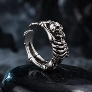 Könnte beinhalten: Ein silberfarbener Metallring mit einem detaillierten Skelett-Design. Die Ringschiene wird durch die Skelettstruktur gebildet, wobei der Schädel oben positioniert ist und der Brustkorb sowie die Armknochen sich zu einem offenen, verstellbaren Ring formen.