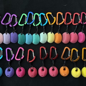 Peut inclure: Une collection de porte-clés composée de mousquetons colorés attachés à de petites prises d'escalade texturées de formes et de teintes variées. L'ensemble comprend des prises en forme de croissant et circulaires en rose, violet, bleu, vert, jaune et orange.