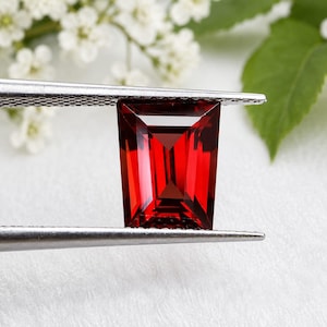 May include: A vibrant, deep red gemstone with a rectangular emerald cut, held securely by a pair of silver-toned metal tweezers. The stone features sharp, clean facets that reflect light, creating a clear, transparent appearance with intense color.