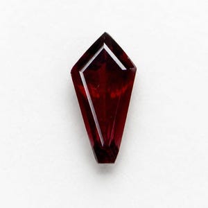 Può includere: Una gemma rosso scuro a forma di aquilone con una superficie sfaccettata. La pietra presenta una parte superiore appuntita e un bordo inferiore piatto, mostrando una qualità traslucida con riflessi interni e bordi geometrici netti.