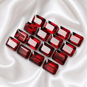 Puede incluir: Una colección de quince piedras preciosas rectangulares de color rojo intenso, talladas en facetas y dispuestas en cuadrícula. Cada piedra presenta un corte esmeralda clásico con esquinas afiladas y superficies reflectantes, mostrando un tono carmesí.