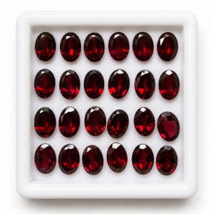 Pode incluir: Uma caixa de exposição quadrada branca contendo vinte e quatro pedras preciosas vermelho-escuras com lapidação oval facetada, dispostas em quatro filas de seis. Cada gema apresenta uma superfície polida com múltiplas facetas que refletem a luz.