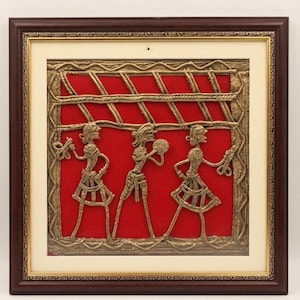 Può includere: Un'opera d'arte murale quadrata che presenta tre figure metalliche color oro in rilievo su uno sfondo di tessuto rosso vibrante. Le figure sono incorniciate da un bordo in legno marrone scuro con una sottile finitura dorata e un passe-partout color crema.