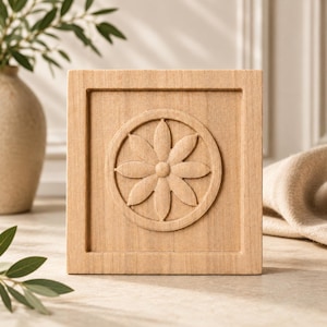 Puede incluir: Un bloque de madera cuadrado de color claro que presenta un marco circular tallado que contiene un diseño de flor de ocho pétalos. La veta de la madera es visible en toda la superficie plana y el motivo floral tallado está centrado.