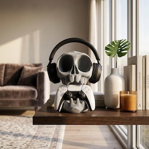 Puede incluir: Un soporte para mando de juegos y auriculares con forma de calavera gris. La calavera tiene cuencas oculares grandes y vacías y lleva auriculares negros. Un mando de juego inalámbrico blanco y negro se sostiene en las manos de la base.