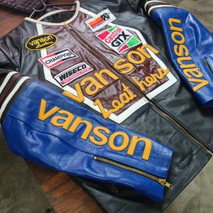 May include: A black, blue, and maroon leather motorcycle jacket featuring yellow Vanson lettering on the sleeves and chest. The jacket includes various patches such as Castrol GTX, Champion, Wiseco, and Vanson Leathers, along with a small American flag patch.
