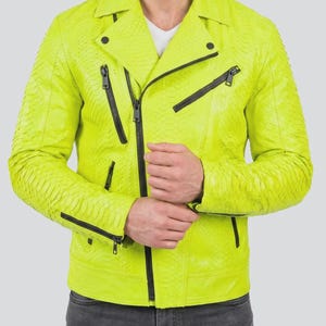 Puede incluir: Una chaqueta de moto de color amarillo neón con un patrón texturizado de piel de serpiente, un cuello ancho con muescas y herrajes negros que incluyen una cremallera frontal diagonal, dos bolsillos en el pecho y puños con cremallera, sobre vaqueros gris oscuro.