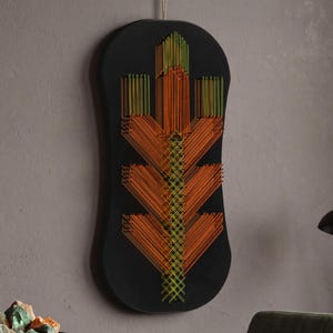 Puede incluir: Una pieza de arte mural negra de forma ovalada vertical con un diseño de arte con hilos. El patrón está creado con hilos naranjas y amarillos dispuestos en una estructura geométrica en forma de hoja que se extiende de abajo hacia arriba.