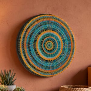 Pode incluir: Uma peça de arte circular com fios sobre uma base escura, apresentando anéis concêntricos de fios em tons de laranja, amarelo e azul-petróleo. O design é criado enrolando fios em torno de pequenos pinos metálicos na borda da moldura.