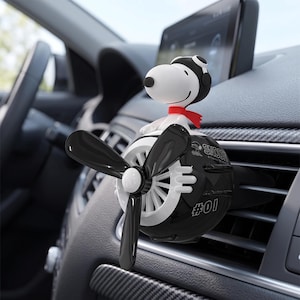 May include: A black and white car air freshener shaped like a vintage airplane with a Snoopy figure wearing a pilot hat and red scarf sitting on top. The propeller is black, and the side of the plane body features the text Snoopy and the number #01.