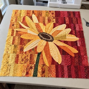 May include: A square quilt block featuring a large, central sunflower appliqué with yellow and orange petals and a dark brown center. The background is composed of vertical fabric strips in a gradient of yellow, orange, and deep red tones.