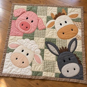 May include: A square patchwork quilt featuring four appliqued farm animal faces: a pink pig, a tan and white cow, a white sheep, and a gray donkey. The background consists of a checkered pattern in shades of cream, sage green, and tan.