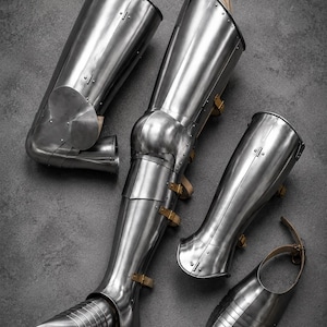 May include: A set of polished steel plate armor for the legs, including greaves, poleyns, and sabatons. The pieces feature articulated joints, metal rivets, and tan leather straps with buckles for secure fastening, showcasing a metallic silver finish.