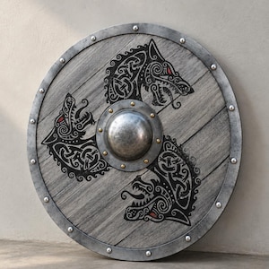 Peut inclure: Un bouclier circulaire en bois à la finition grise patinée, doté d'un umbo métallique central fixé par des rivets en laiton. Trois têtes de loup noires aux yeux rouges et aux motifs entrelacés sont disposées en triangle autour du centre.