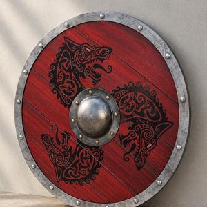 May include: A circular wooden shield with a red stained finish and a metallic rim secured by rivets. The center features a domed steel boss. Three black wolf heads with intricate knotwork patterns are arranged in a triangular formation.