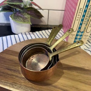 May include: A set of three nested measuring cups with round copper bowls and long, angled brass handles. The cups are stacked inside one another, displaying a metallic finish with visible signs of use and a warm, reflective copper and brass tone.