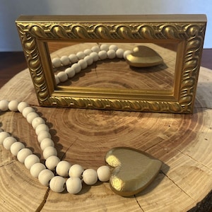May include: A rectangular mirror with an ornate gold frame featuring a repeating leaf pattern. A strand of round, unfinished wooden beads is draped in front of the mirror, ending in a smooth, solid gold-colored heart-shaped ornament.